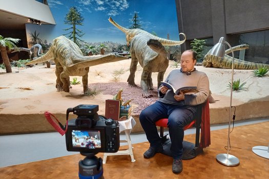 Mann sitzt vor Saurier Diorama und liest vor. Er wird dabei von einer Kamera gefilmt.