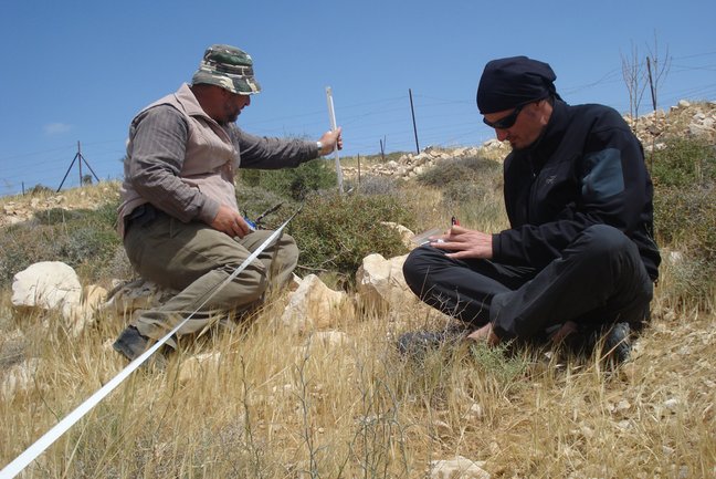 Zwei Wissenschaftler bei der Feldarbeit untersuchen Pflanzen in einer trockenen Steppenlandschaft.