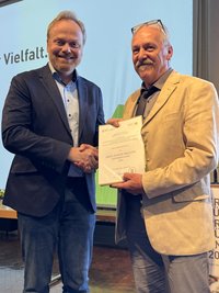 Lars Krogmann hat Axel Hofmann die Urkunde über den Erhalt des Ernst-Jünger-Preises überreicht. Lars Krogmann steht links im Bild, lächelt in die Kamera und schüttelt Axel Hofmann die Hand. Axel Hofmann steht rechts im Bild hält in seiner linken Hand die Urkunde und schüttelt mit seiner Rechten die Hand von Lars Krogmann.