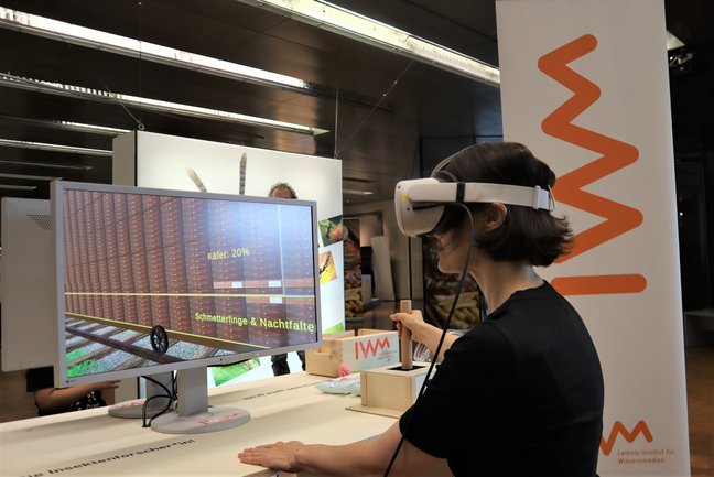 Person mit VR-Brille bedient einen Joystick vor einem Monitor.
