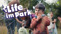 Junge mit Lupe schaut in Kamera, im Hintergrund mehrere Personen und dazu der Schriftzug Life im Park