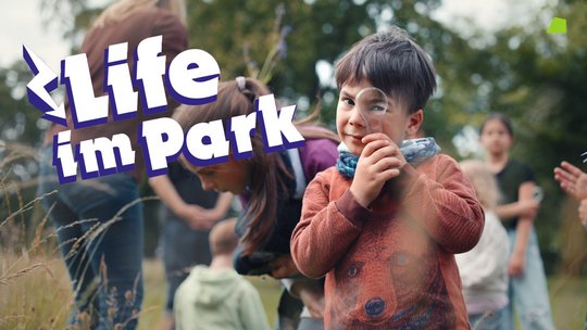 Junge mit Lupe schaut in Kamera, im Hintergrund mehrere Personen und dazu der Schriftzug Life im Park