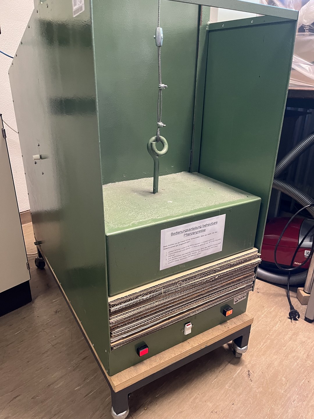 Eine professionelle Pflanzenpresse. In einem schrankartigen Gerüst aus Metall liegt ein Gewicht auf einem Stapel Wellpappe. 