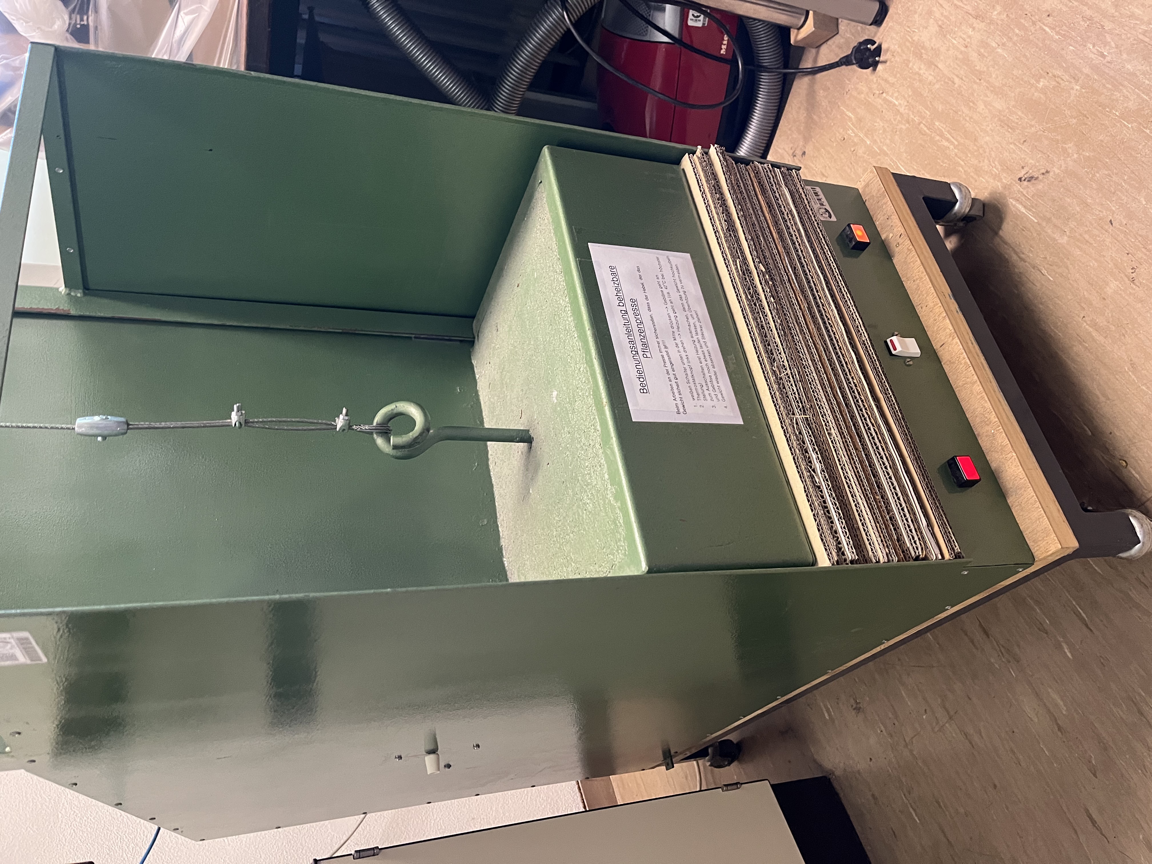 Eine professionelle Pflanzenpresse. In einem schrankartigen Gerüst aus Metall liegt ein Gewicht auf einem Stapel Wellpappe.