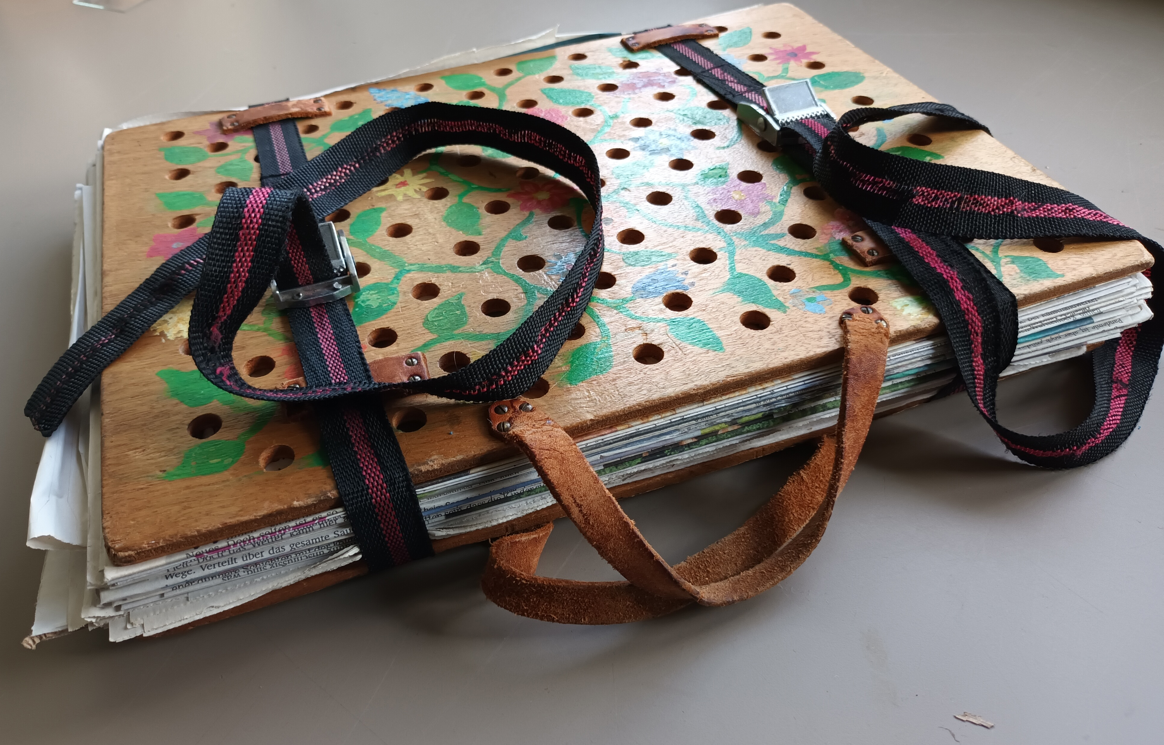 Eine Pflanzenpresse. Zwischen zwei rechteckigen Holzplatten ist ein Stapel gefalteter Zeitungen zu erkennen. Die Holzplatten und Zeitungen werden durch zwei Spanngurte zusammengehalten. Die obere Holzplatte hat viele kleine runde Löcher und ist mit einem grünen Pflanzenmuster verziehrt.