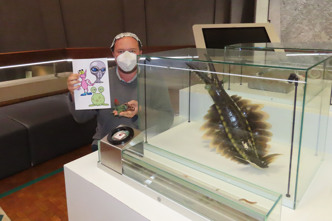Mann sitzt neben einer Vitrine mit der Rekonstruktion eines urzeitlichen Meereslebewesen und hält Abbildungen von Aliens.