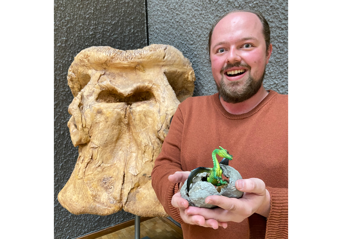 Museumspädagoge Malte Aurich steht neben dem Schädel eines Mammuts und hält ein zerbrochenes Ei, aus dem ein Spielzeugdrache herausschaut.